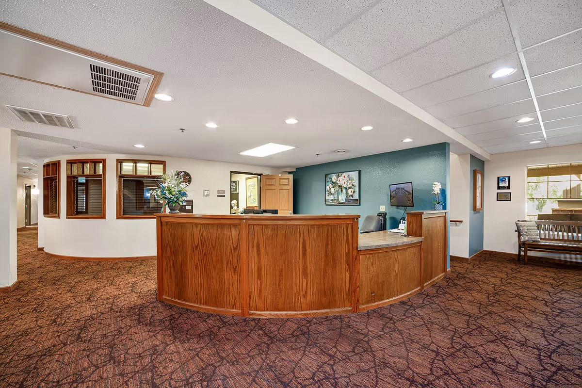 Reception area with a curved wooden front desk, a computer monitor, and a vase with flowers. The walls are painted white and teal, with framed artwork and windows behind the desk. There is a bench with cushions near a window on the right side and patterned carpet on the floor.