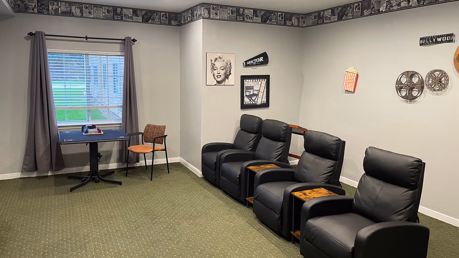 A room with four black leather recliner chairs with wooden armrest trays lined up against a light gray wall decorated with movie-themed wall art including film reels, a popcorn box, and a Marilyn Monroe portrait. There is a window with gray curtains and a small table with a single chair near it. The room has green carpet and a movie-themed border along the top of the walls.