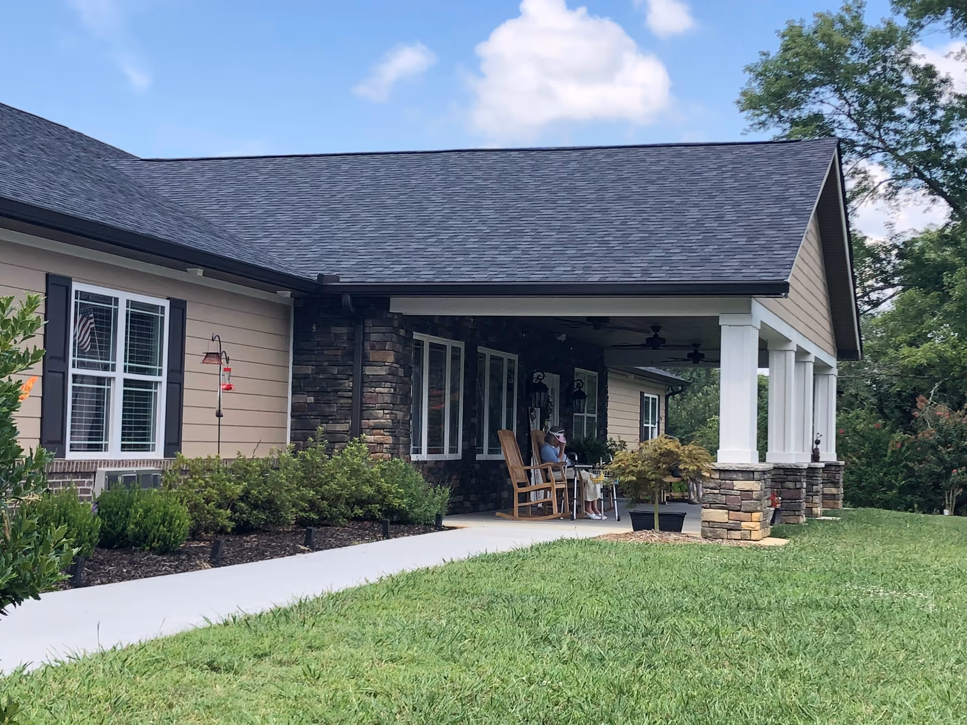 Exterior view of a single-story building with beige siding and stone accents. There is a covered porch with white columns and two wooden rocking chairs, one occupied by a person. The porch overlooks a well-maintained grassy lawn and garden beds with shrubs. The sky is partly cloudy.