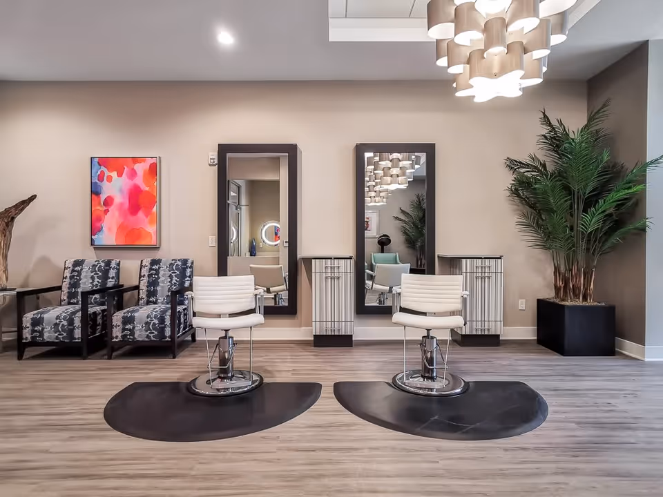 Interior view of a senior living facility's salon area with two white salon chairs in front of two large mirrors mounted on the wall. There are two patterned armchairs to the left, a colorful abstract painting on the wall, and a large potted plant to the right. The floor is wood, and a modern chandelier hangs from the ceiling.