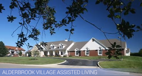 Single-story brick-and-siding assisted living building with a curved driveway, lawn, and trees under a clear blue sky.