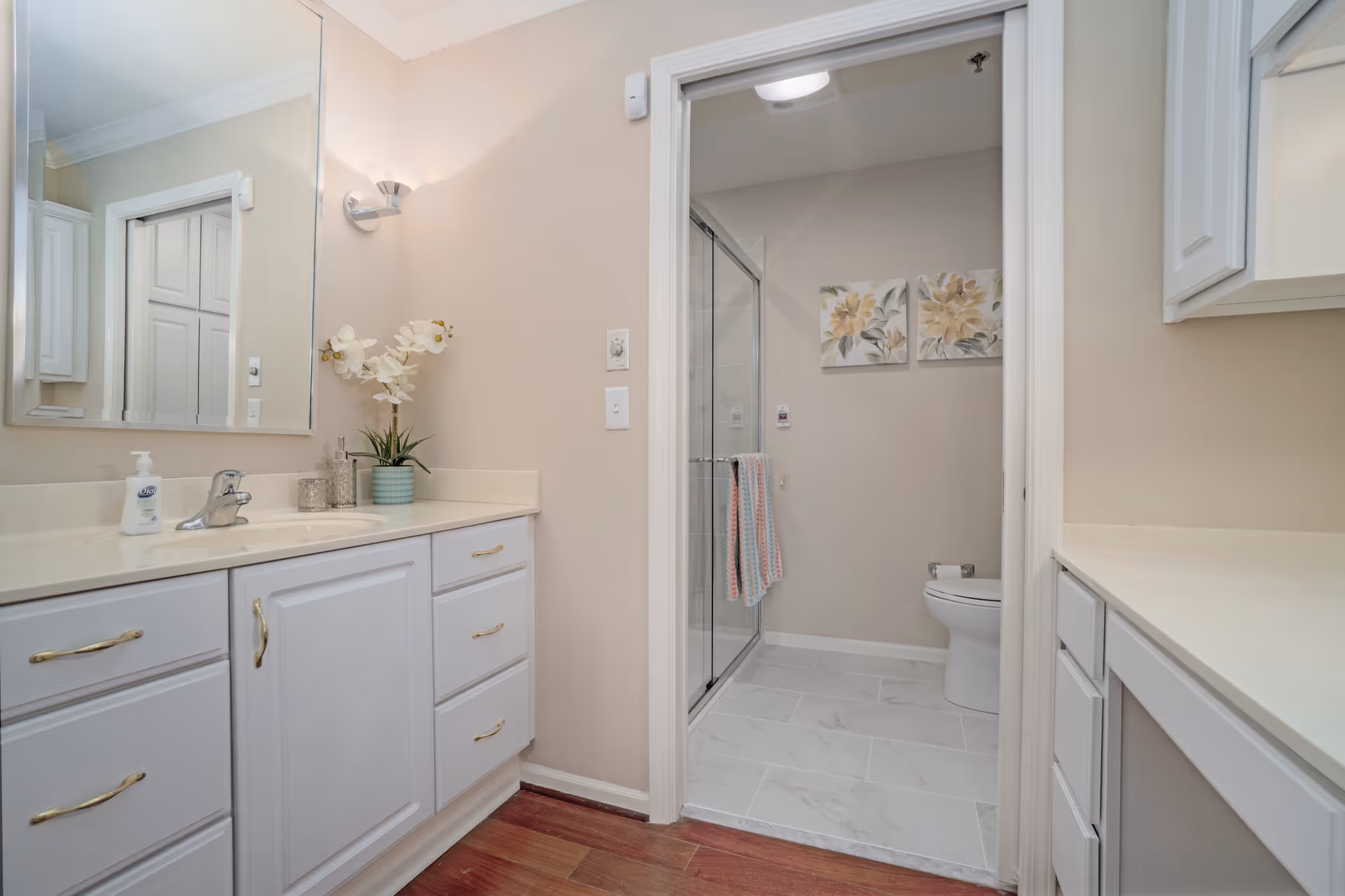 Bright bathroom with white vanity and cabinets, a sink and mirror, and a separate toilet with a glass-enclosed shower.