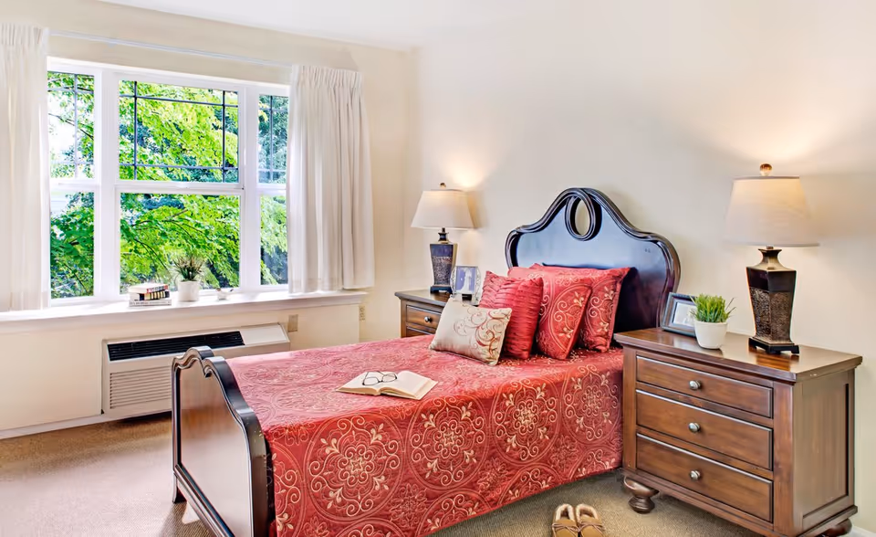 Sunlit bedroom with a wooden bed dressed in red patterned bedding, matching nightstands and lamps beside a large window.