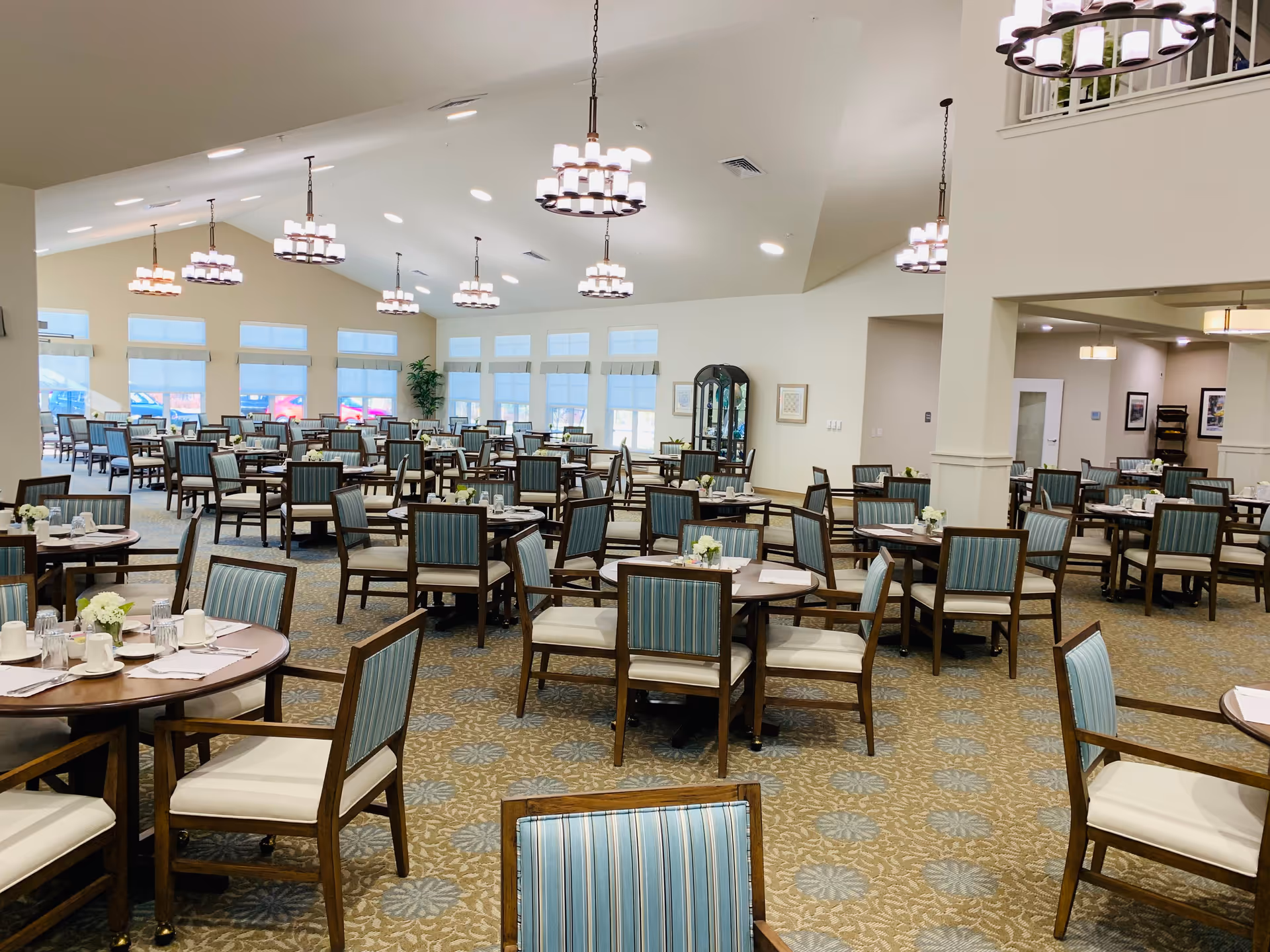 Spacious bright dining room with many round tables and striped chairs set for meals under chandeliers.