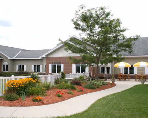 Outdoor patio area at North Terrace Assisted Living with a paved walkway, landscaped garden beds with flowers and shrubs, a tree, and tables with yellow and white striped umbrellas next to the building.