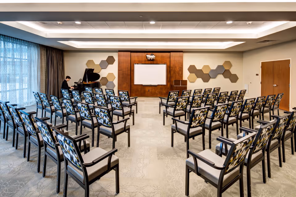 A conference or presentation room with rows of patterned chairs facing a whiteboard and a wooden panel wall. A person is playing a grand piano on the left side near large windows with sheer curtains. The room has modern ceiling lighting and hexagonal wall decorations.