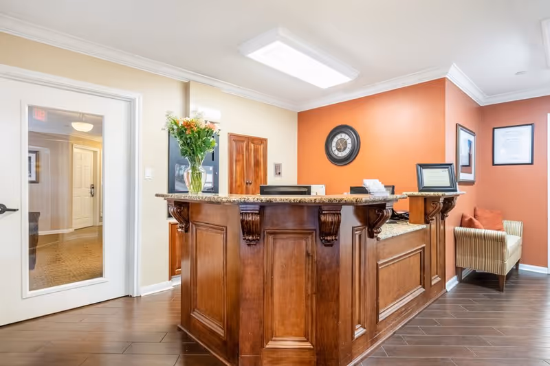 A warm lobby reception area with a polished wooden front desk, granite countertop, orange accent wall, wall clock, and a striped armchair.