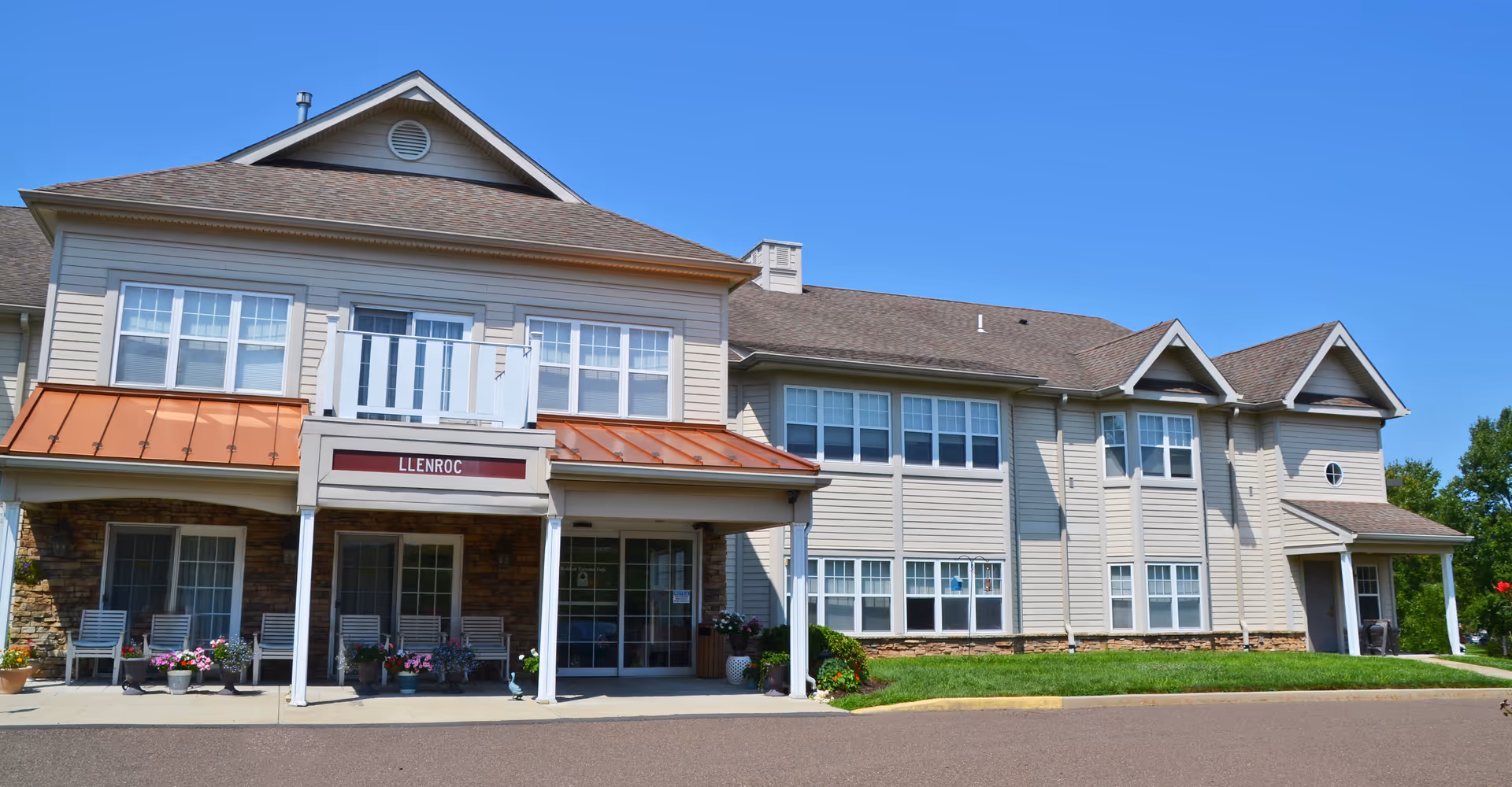 Exterior view of a two-story senior living facility building with beige siding, multiple windows, a covered entrance with white pillars, and a small balcony above the entrance. There are potted plants and chairs near the entrance, and the sky is clear and blue.