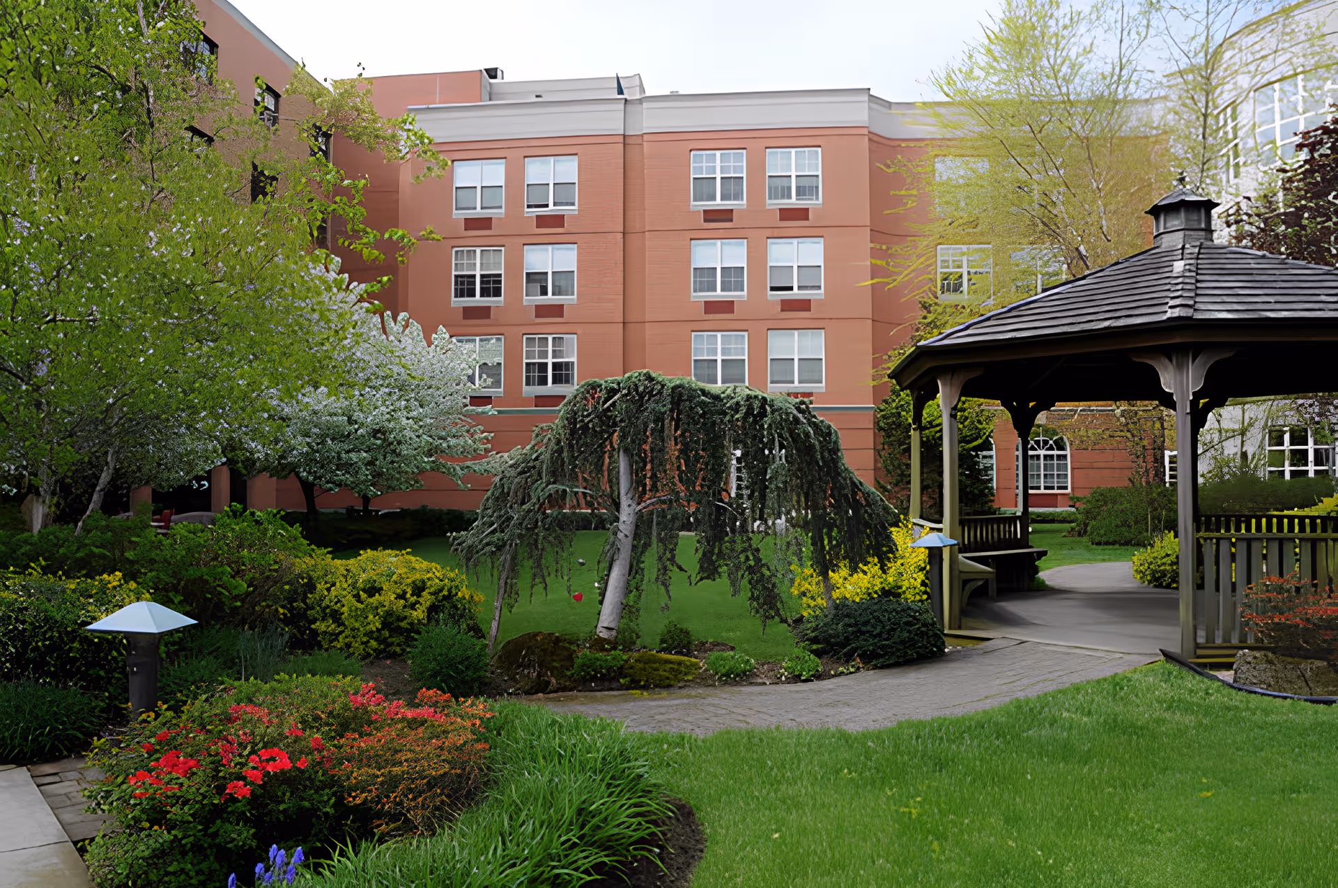 A landscaped garden area with green grass, various shrubs, and flowering plants in front of a multi-story brick building. There is a wooden gazebo with a shingled roof on the right side of the image, and a paved walkway leading through the garden.