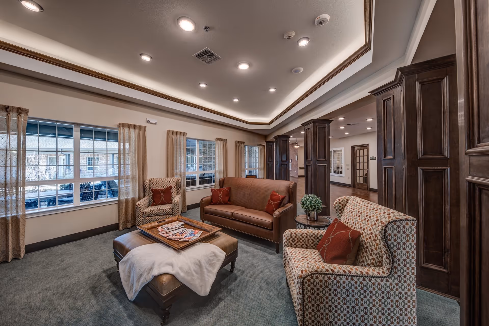 A cozy assisted living facility common area with a brown leather sofa, two patterned armchairs with red cushions, a large ottoman with a white throw blanket and a tray of magazines, and large windows with beige curtains letting in natural light. The room features dark wood columns and trim, recessed ceiling lights, and a carpeted floor.