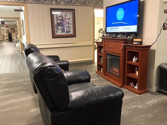 Hallway seating area with black leather recliners facing a wall-mounted TV above a wooden electric fireplace in a senior living facility corridor.