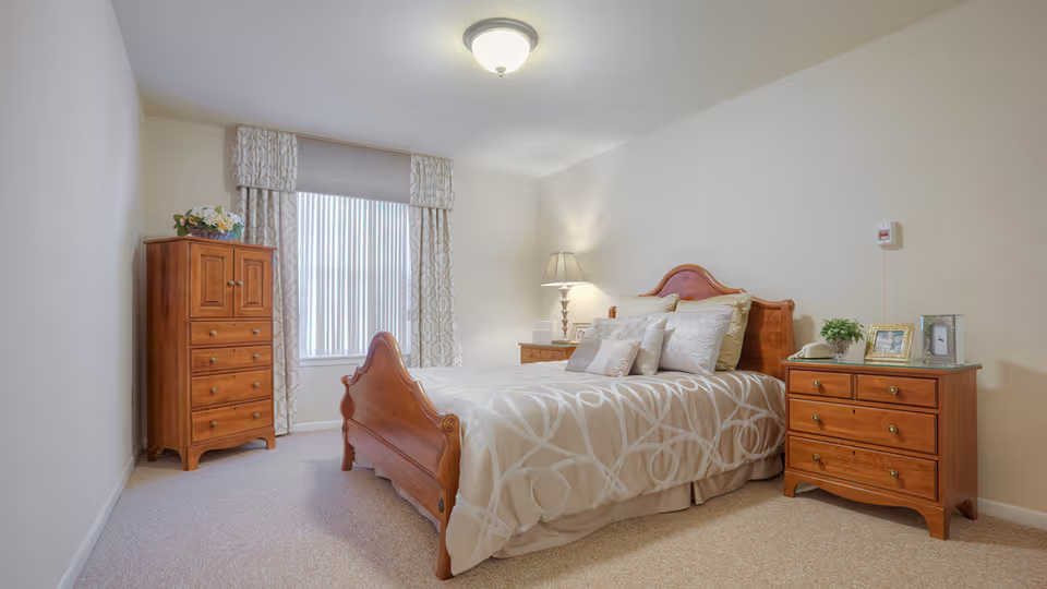 A cozy bedroom in a senior living facility featuring a wooden bed with beige and white patterned bedding, two wooden nightstands with lamps and framed photos, a wooden chest of drawers, and a window with patterned curtains allowing natural light to enter.