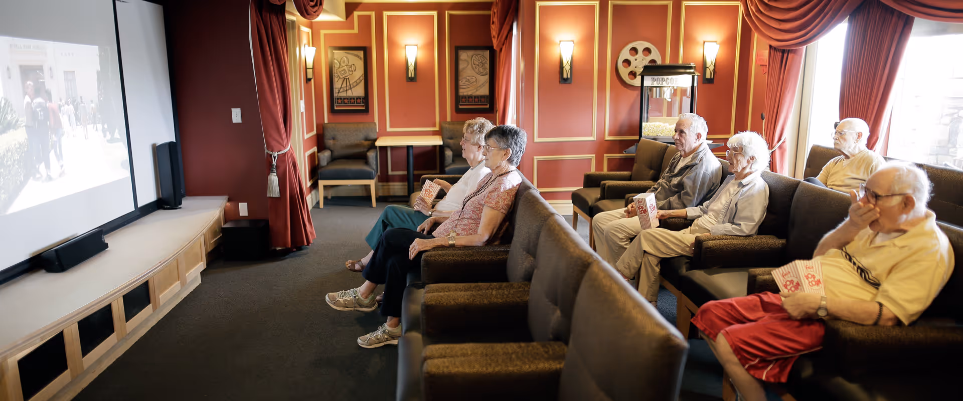 A group of elderly people seated in a small theater room watching a movie on a large screen. They are sitting in comfortable chairs with popcorn, and the room has red walls with decorative paneling and wall sconces. A popcorn machine is visible in the background near a window with red curtains.
