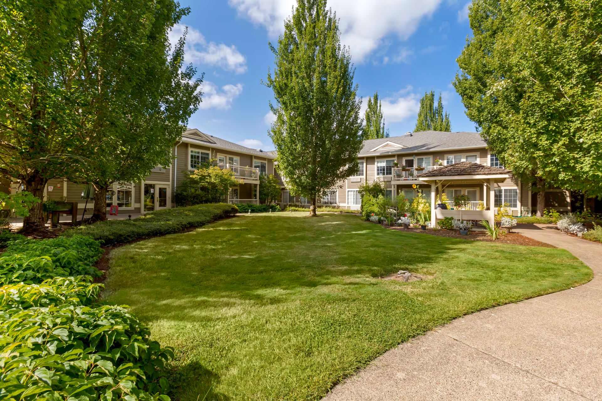 A well-maintained outdoor courtyard area at a senior living facility with green grass, trees, shrubs, and a paved walkway. Two-story buildings with balconies and windows surround the courtyard under a partly cloudy blue sky.