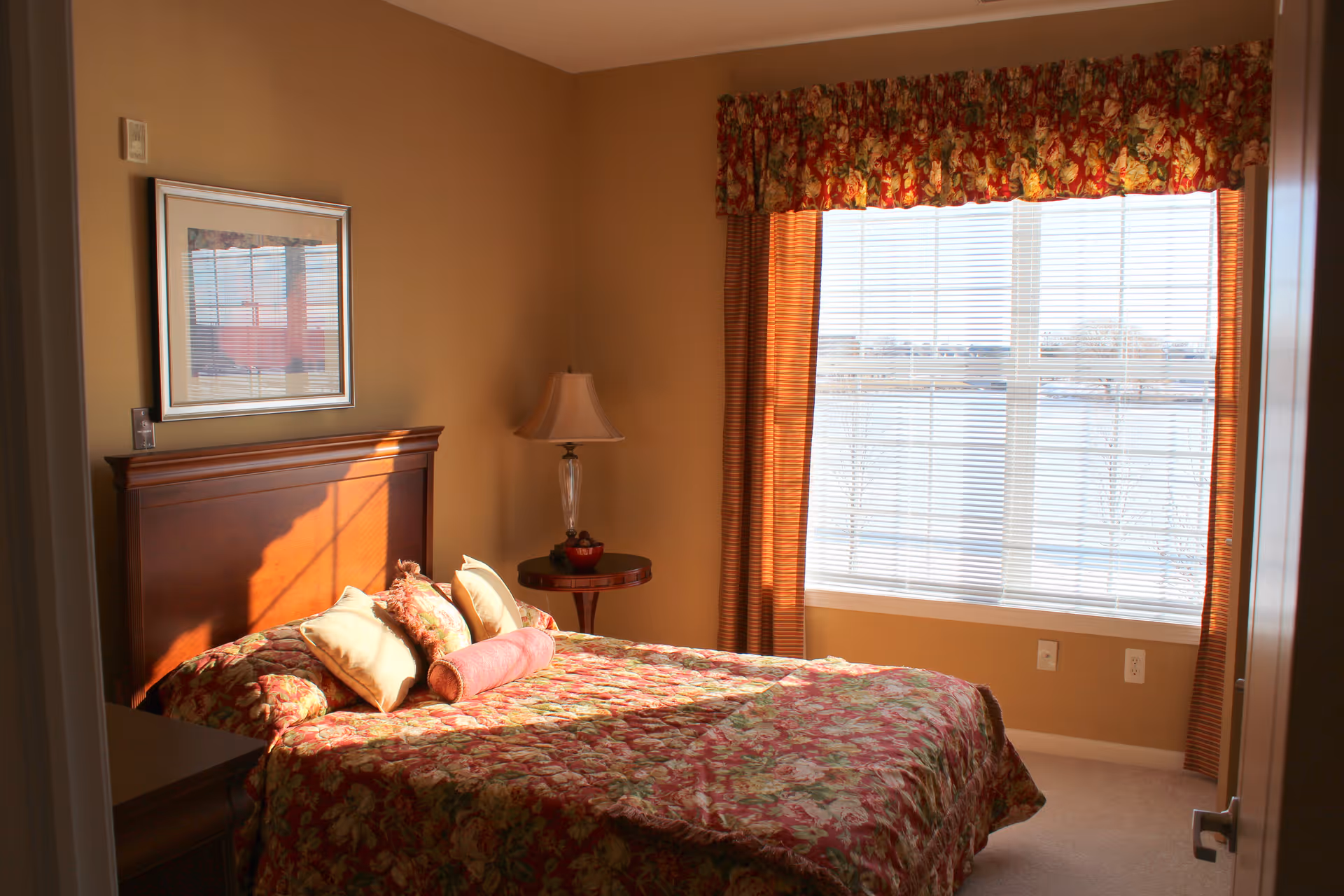 A cozy bedroom with a wooden headboard bed covered in a floral patterned bedspread and several pillows. There is a round wooden side table with a lamp and a decorative bowl next to the bed. A large window with blinds and floral valance allows natural light to fill the room.