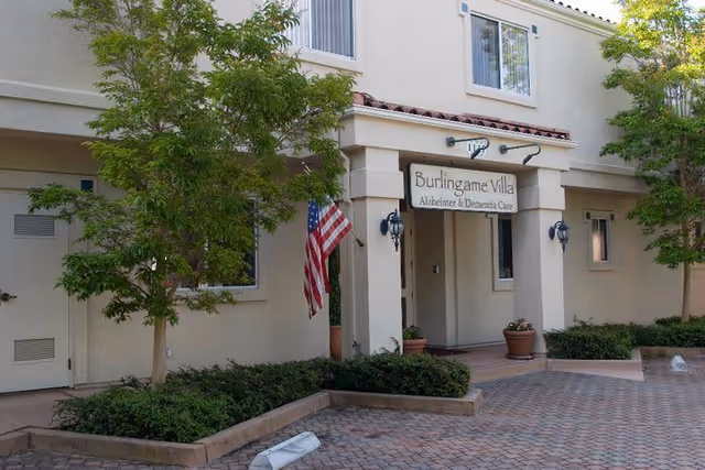 Exterior view of Burlingame Villa, a beige two-story building with a sign reading 'Burlingame Villa Alzheimer & Dementia Care' above the entrance. There are two small trees and shrubs planted in front, an American flag mounted near the entrance, and a paved walkway leading to the door.