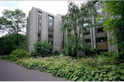 Exterior view of a multi-story building surrounded by trees and lush greenery, with a paved pathway leading alongside the garden area.
