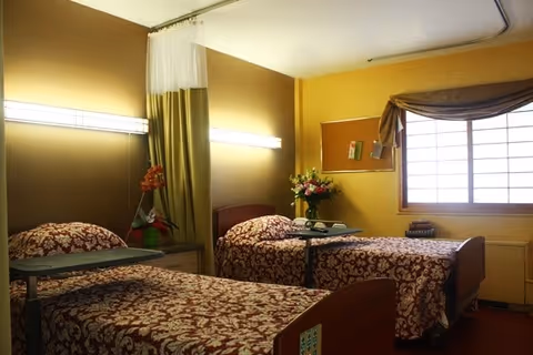 A room with two hospital-style beds covered with red and white patterned bedspreads. Each bed has an overbed table, and there is a bouquet of flowers on a small table between the beds. The walls are painted yellow, and there is a window with a curtain on the right side. A bulletin board is mounted on the wall above the flowers.