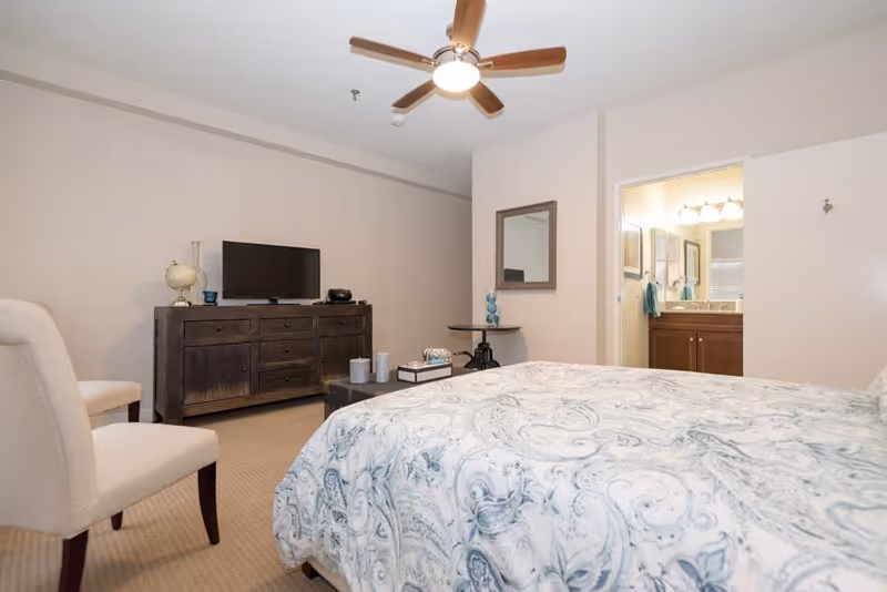 A bedroom with a bed covered in a white and blue patterned comforter, two beige chairs, a dark wooden dresser with a TV and decorative items on top, a ceiling fan with lights, and an open door leading to a bathroom with a vanity and mirror.