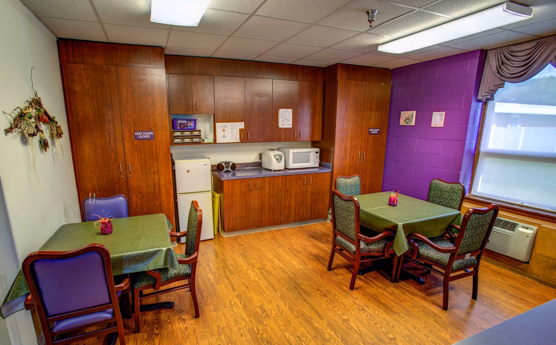 Small communal dining room with two tables and chairs and a kitchenette with wooden cabinets, a microwave and mini fridge.