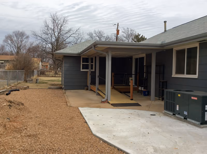 Exterior view of a gray building with a covered porch area featuring a wooden ramp with handrails. The ground is covered with gravel and concrete, and there is an air conditioning unit on the right side. Leafless trees and a chain-link fence are visible in the background under a cloudy sky.