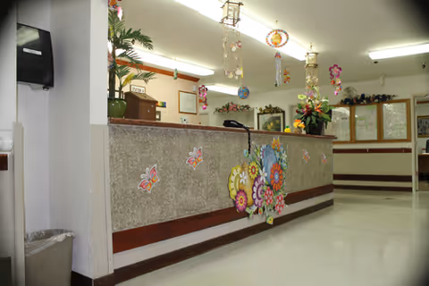 Reception area of a medical center convalescent hospital with a decorated front desk featuring colorful flower and butterfly stickers. The desk has a telephone and a vase with flowers on top. The room is well-lit with fluorescent lights and has white walls and floors. There are hanging decorations from the ceiling and a paper towel dispenser mounted on the wall to the left.