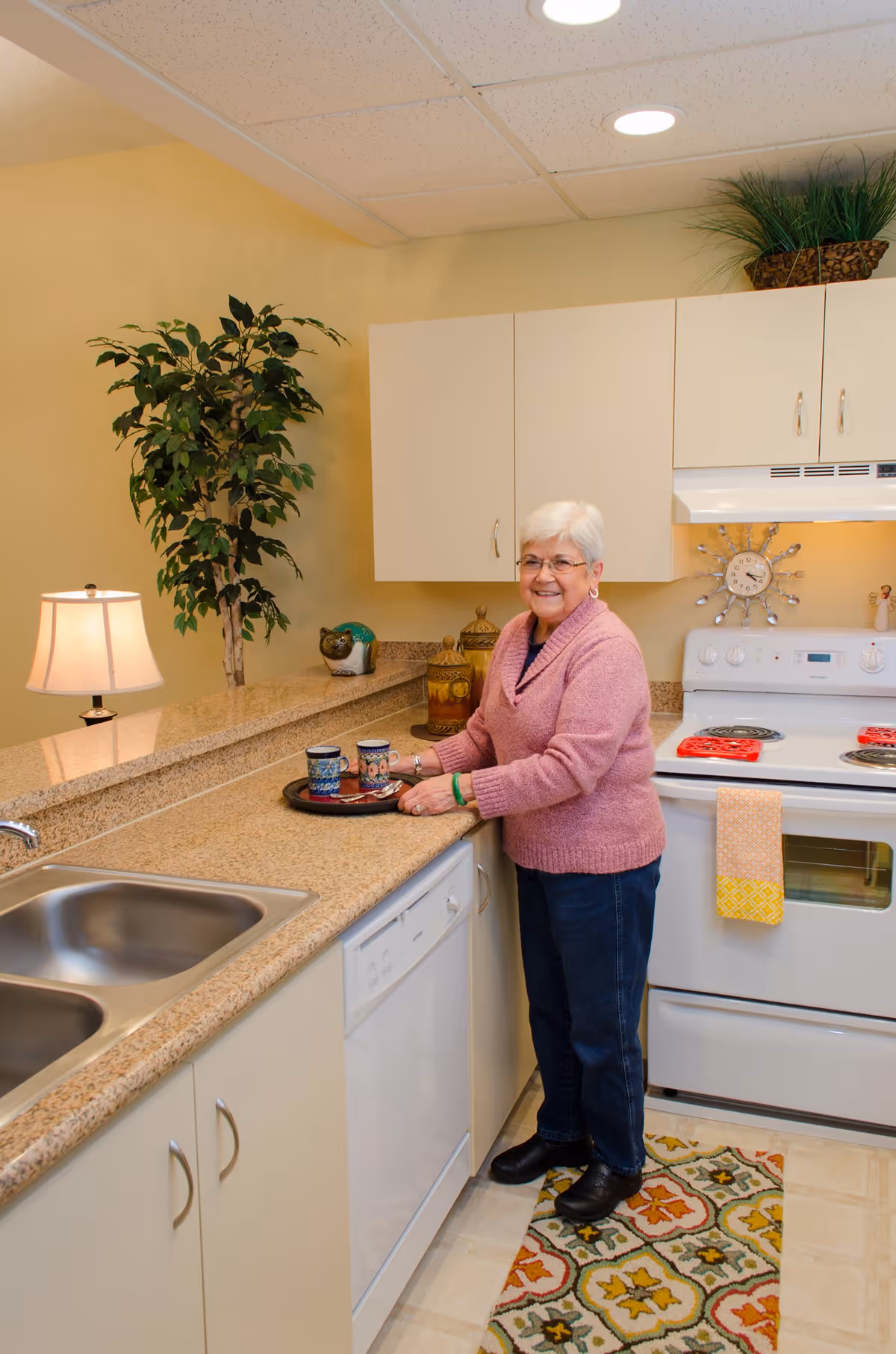 An elderly woman with white hair and glasses stands in a kitchen, smiling and holding a tray with two colorful mugs. The kitchen features beige countertops, white cabinets, a white stove with a yellow and orange towel hanging on the handle, a dishwasher, a stainless steel sink, a decorative plant, and a patterned rug on the floor.