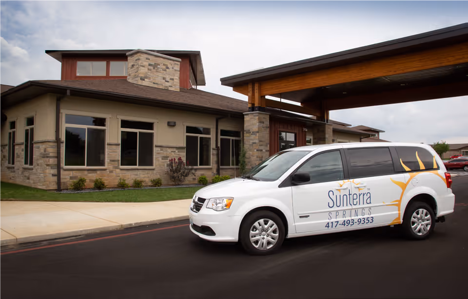 Exterior view of Sunterra Springs Springfield facility with a white Sunterra Springs van parked in front under a covered entrance. The building has stone and beige walls with multiple windows and a chimney.