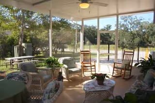 A screened-in porch with wicker furniture including chairs and a table with a potted plant, two wooden rocking chairs, ceiling fan, and a view of trees and a fenced yard outside.