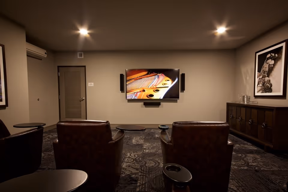A cozy media room with three brown leather chairs facing a wall-mounted flat-screen TV displaying a close-up image of a piano's inside. The room has dim lighting, patterned carpet, a wooden cabinet on the right side with glassware, and framed artwork on the walls.