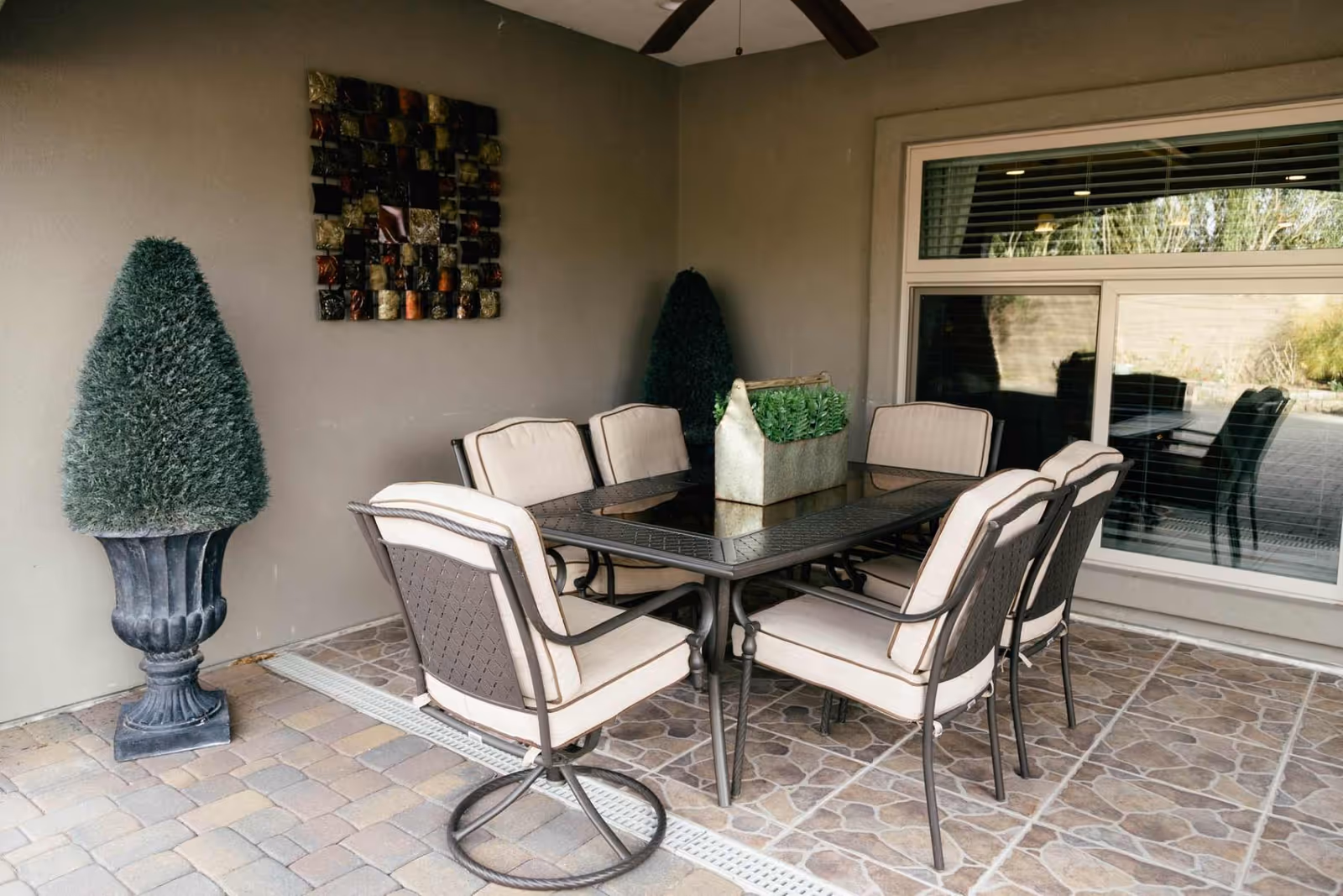 Outdoor covered patio area with a rectangular glass-top table surrounded by six cushioned metal chairs. Two large potted topiary plants are placed against the wall, and a decorative metal wall art piece hangs above. A large window with blinds reflects the outdoor space.