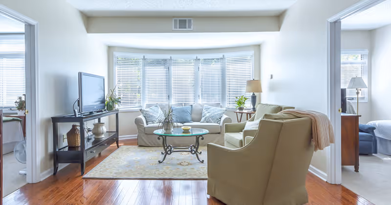 Bright living room with wooden floor, beige sofa and armchair, glass coffee table on a patterned rug, TV on a black stand, and large windows with white blinds letting in natural light. Adjacent rooms with beds are visible on both sides.