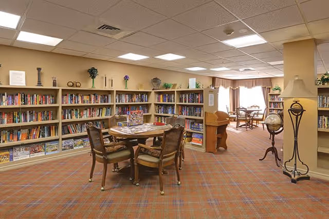 A cozy library room in Regency Oaks Health Center with bookshelves filled with books lining the walls. In the center, there is a round wooden table surrounded by four cushioned chairs. The room has a warm carpet with a plaid pattern, a globe on a stand, a floor lamp, and a window with curtains letting in natural light. Various decorative items are placed on top of the bookshelves.