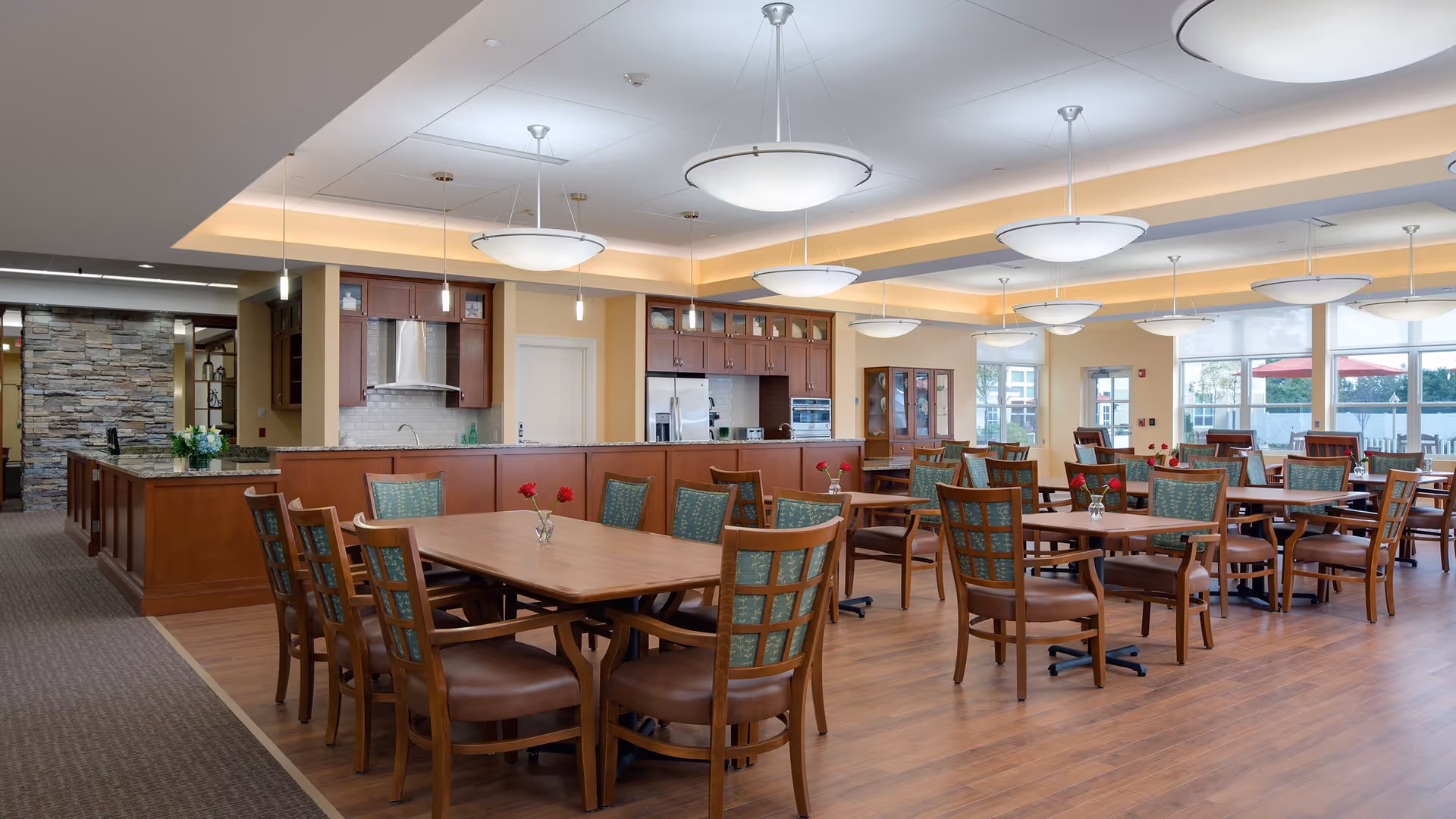 A spacious dining room with multiple wooden tables and chairs, each table decorated with a small vase holding a red rose. The room features a large open kitchen area with wooden cabinets, stainless steel appliances, and a stone accent wall. Large windows allow natural light to fill the space, and modern pendant lights hang from the ceiling.
