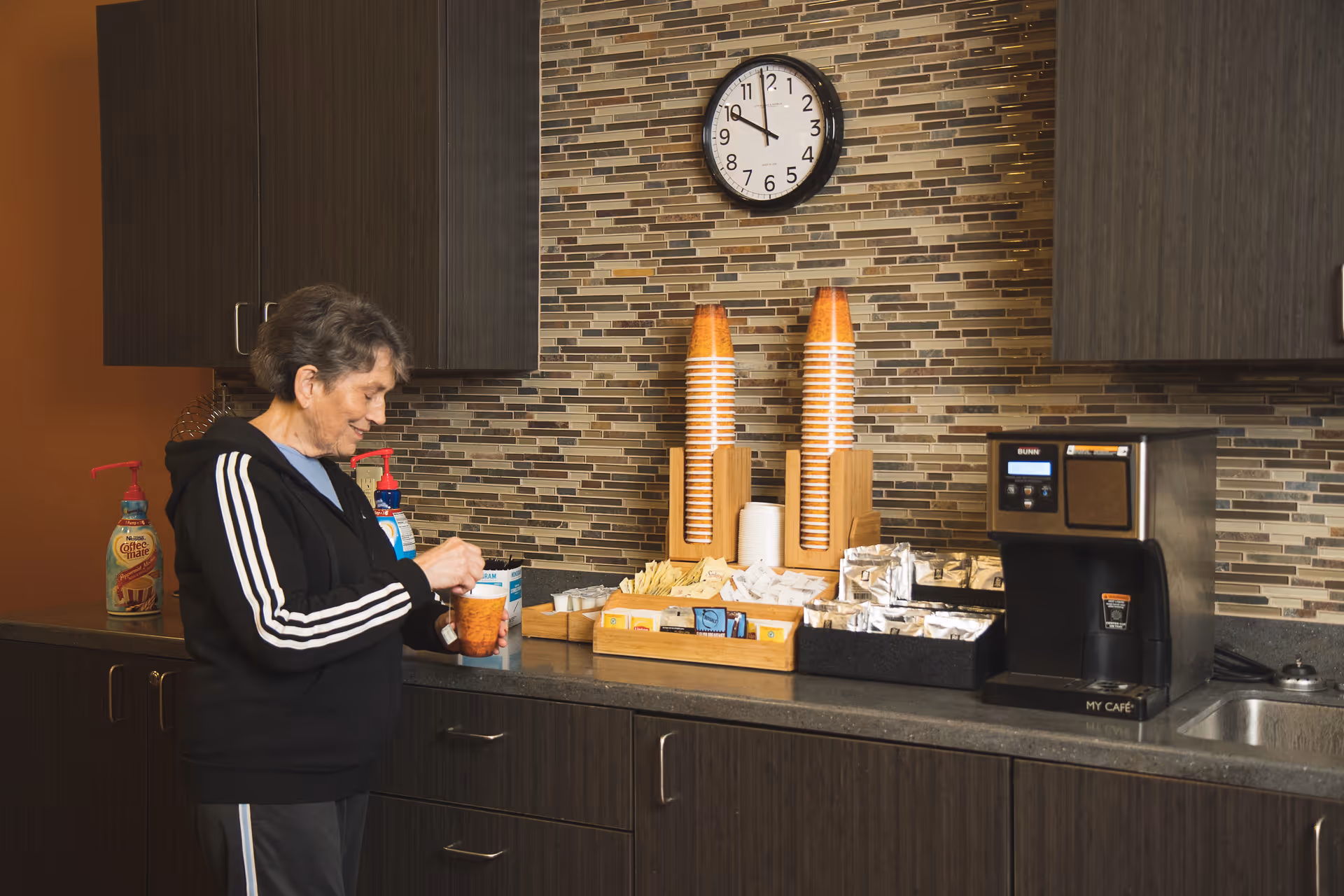 A person preparing a hot drink at a kitchenette coffee station with stacked disposable cups, condiments, and a coffee machine.