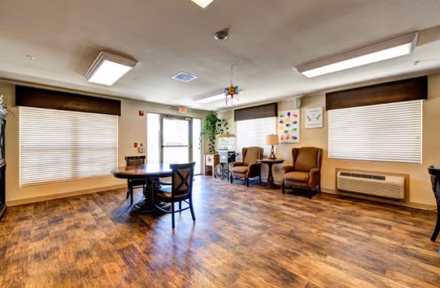 Bright senior living common room with a round table, chairs, armchairs and large windows.