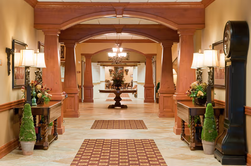 Well-lit elegant interior hallway with wooden columns, side tables, lamps, decorative plants and a central table topped with a floral arrangement.