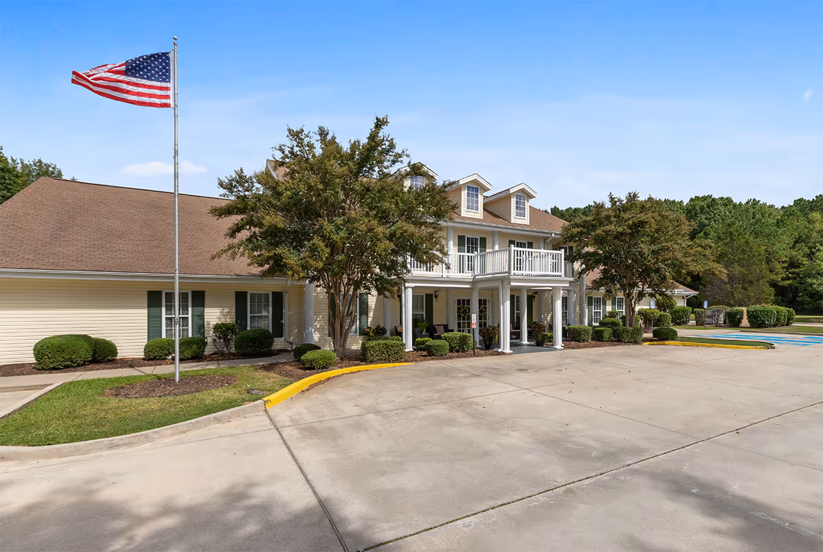 Exterior view of a senior living facility building with beige siding and a brown roof. The building features a covered entrance with white columns and a balcony above it. There are several windows with green shutters, well-maintained bushes, and trees around the building. An American flag is flying on a flagpole near the entrance. The sky is clear and blue.