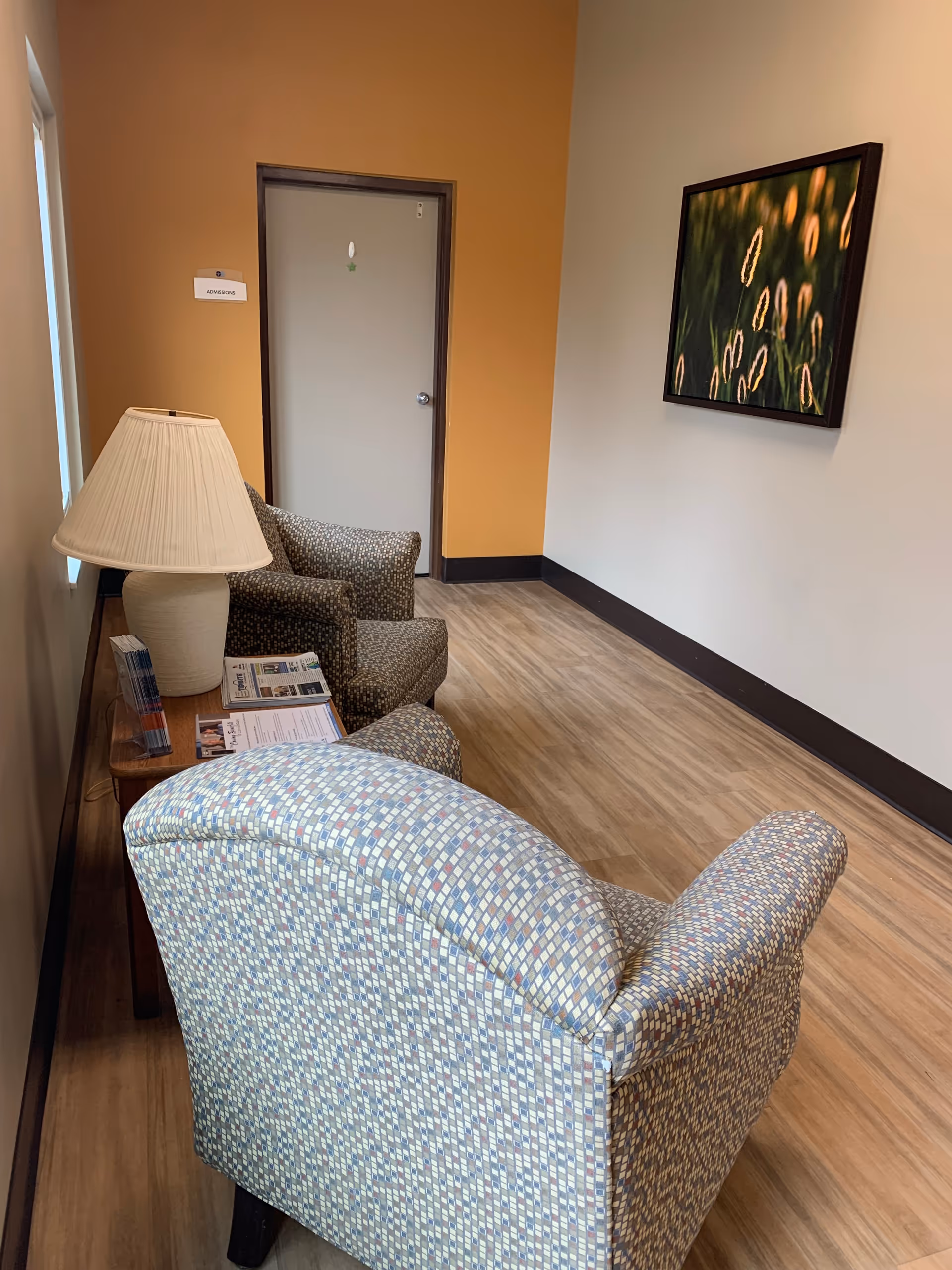 A small waiting area with two patterned armchairs and a wooden side table holding a lamp and some brochures. The walls are painted in beige and orange tones, with a framed picture of plants on the wall. A door labeled 'Admissions' is visible at the end of the hallway.