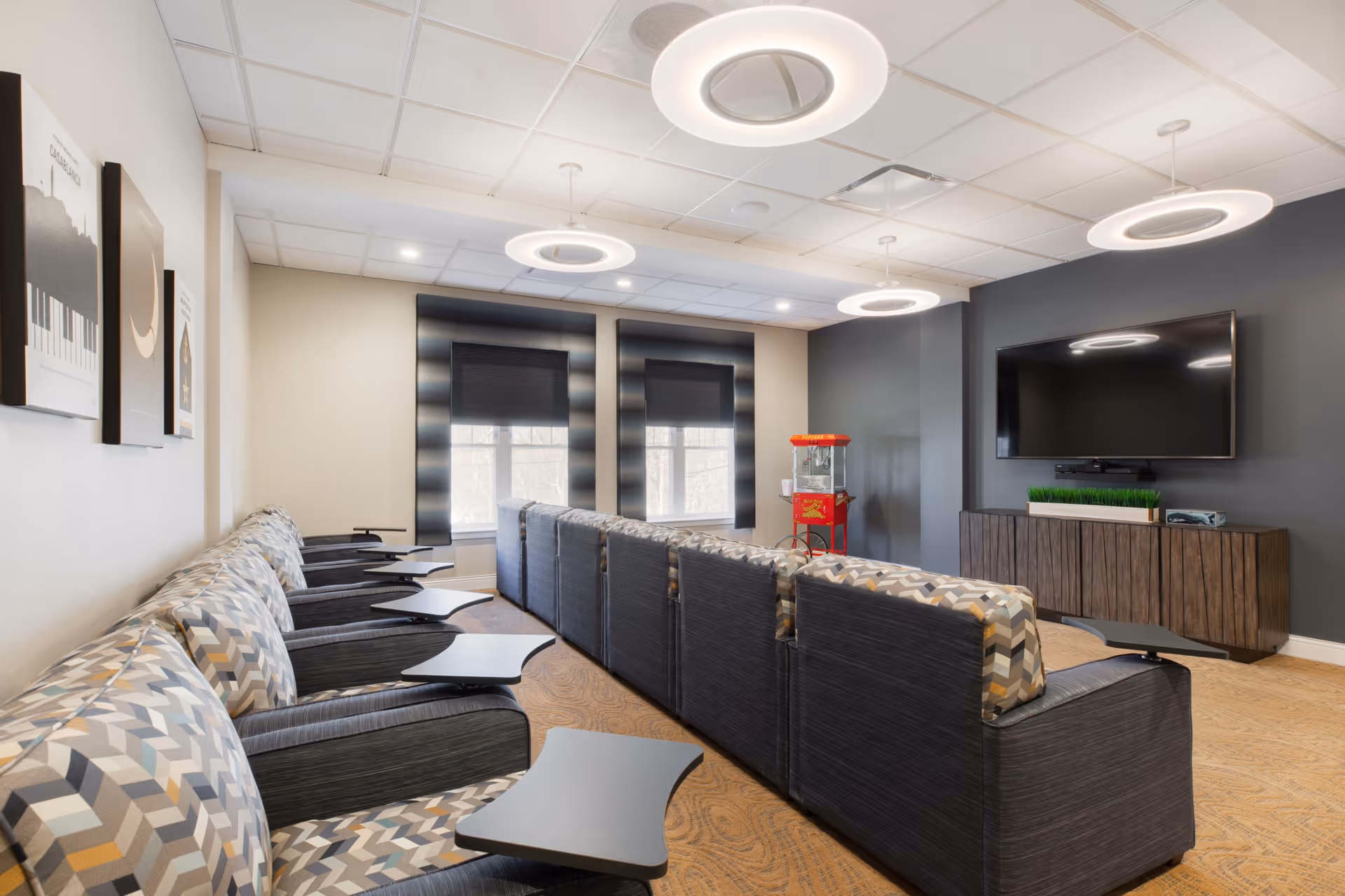 A modern lounge area with two rows of patterned armchairs equipped with individual black tray tables facing a large flat-screen TV mounted on a dark gray accent wall. The room features circular ceiling lights, two windows with black blinds, a wooden cabinet below the TV, and a red popcorn machine in the corner.