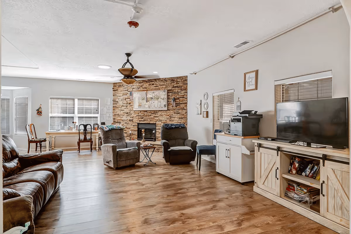 A spacious living room with wooden flooring featuring a brown leather sofa on the left, two gray recliners in the center near a stone fireplace with a painting above it. There is a wooden table with chairs by the window in the background, a TV on a wooden cabinet on the right, and a printer on a white cabinet next to it. The room has white walls and several windows with blinds.
