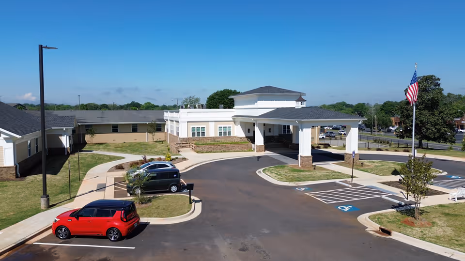 Front exterior of a senior health facility with a covered entrance/porte-cochere, parking lot with cars, landscaped grounds, and an American flag.