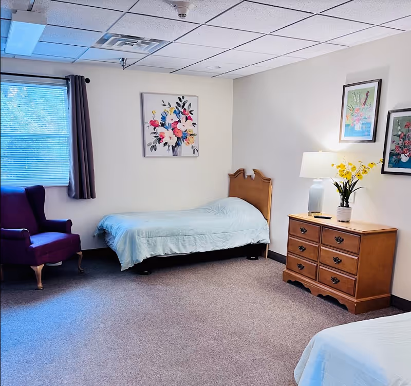 A bedroom in an assisted living facility with two single beds covered in light blue bedding. There is a wooden dresser with a lamp and a vase of yellow flowers on top. The walls are decorated with three framed floral paintings. A purple armchair is positioned near a window with closed blinds and a gray curtain.