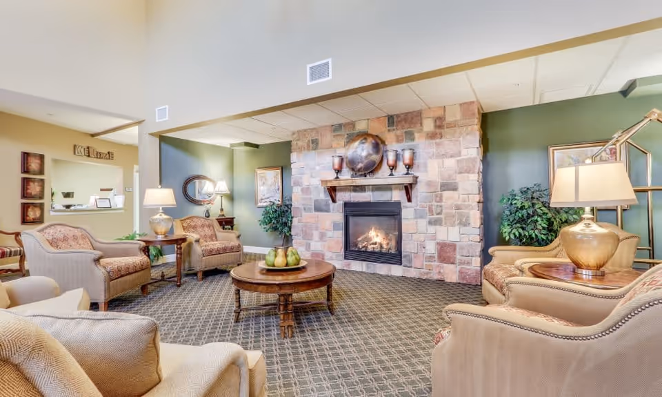 A cozy living room area in a senior living facility featuring a stone fireplace with a fire burning, a round wooden coffee table with decorative pears, several upholstered armchairs and sofas arranged around the fireplace, table lamps, framed artwork on the walls, and green plants adding to the warm and inviting atmosphere.