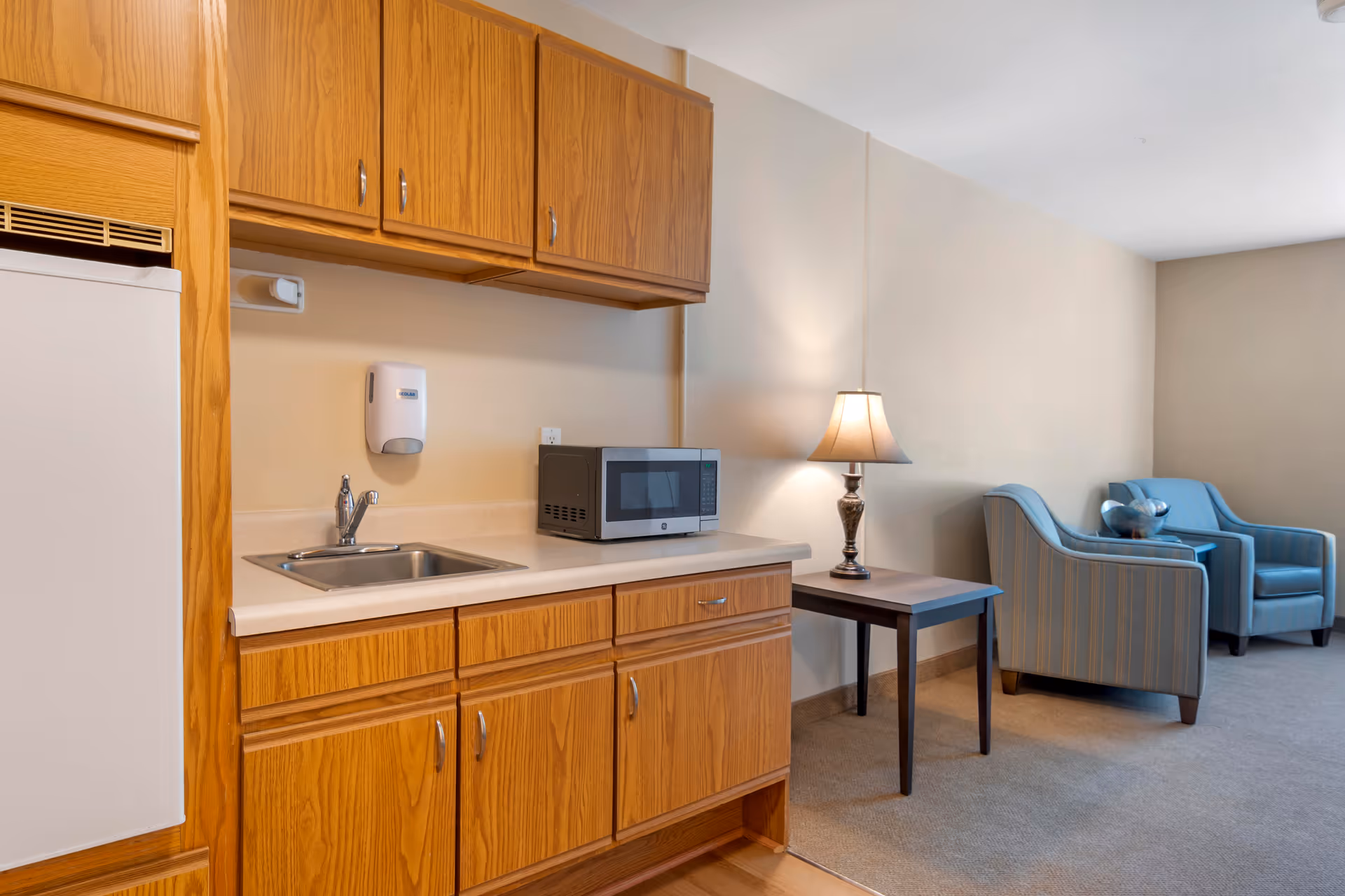 A small kitchenette with wooden cabinets, a sink, a microwave, and a mini refrigerator. Next to the kitchenette is a small wooden side table with a lamp on it. In the background, there are two blue upholstered armchairs with a small table between them, set against a beige wall.