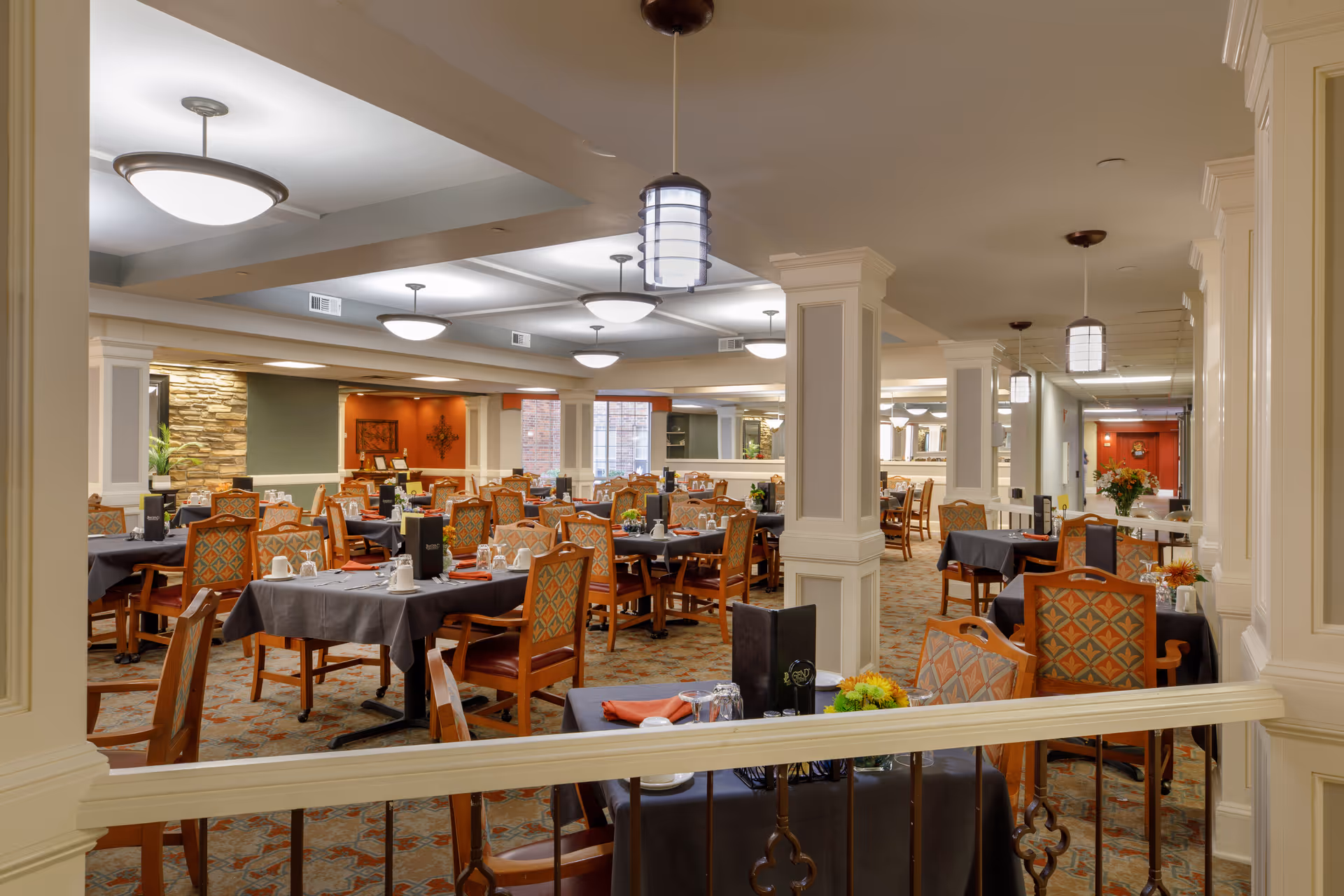 A spacious dining room in Rivermont Independent Living with multiple tables covered in dark tablecloths, each set with cups, glasses, and napkins. The room features patterned chairs, carpeted floors, white columns, and warm lighting from ceiling fixtures. The walls have decorative elements including a stone accent wall and artwork.