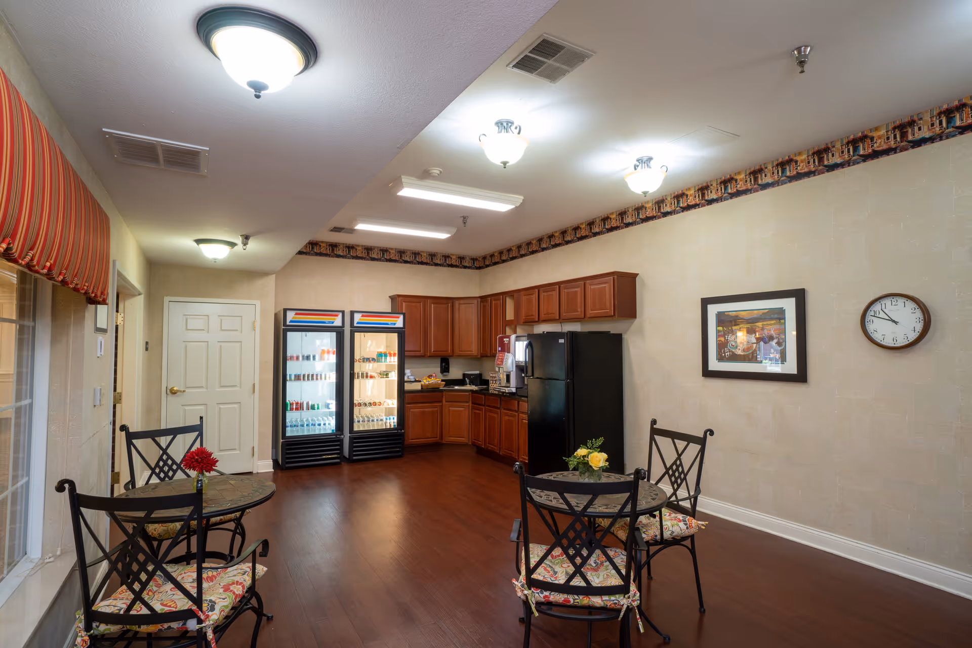 A small dining area with two round tables and four chairs with floral cushions. The room has wooden flooring, beige walls, and a kitchen area with wooden cabinets, a black refrigerator, a coffee machine, and two glass-door refrigerators stocked with beverages. There is a framed picture and a clock on the wall, and a window with a red and beige striped valance.