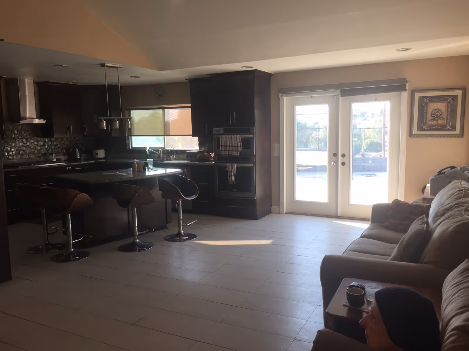 Open-plan kitchen and living area with a dark wood island and bar stools, tiled floor, sofa, and glass French doors letting in light.