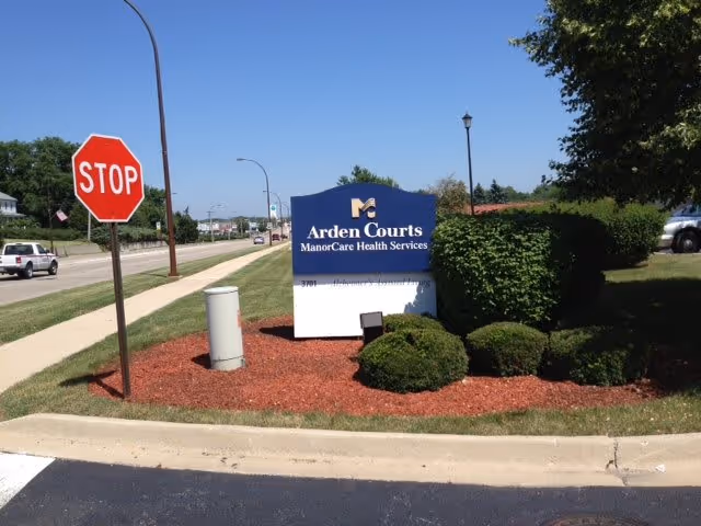 Outdoor view of a landscaped area next to a road with a stop sign and a blue and white sign for Arden Courts ManorCare Health Services surrounded by bushes and mulch.