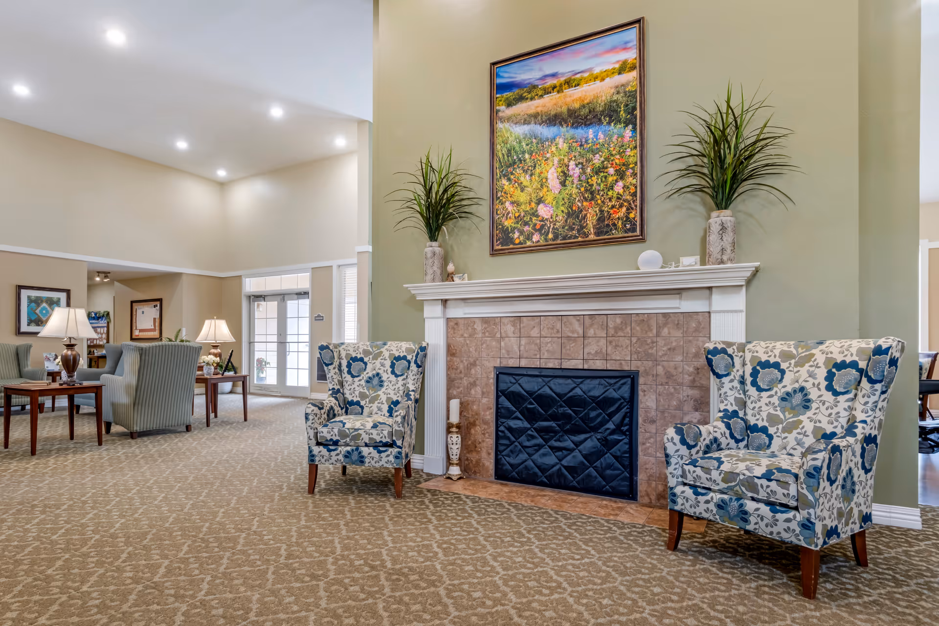 Spacious lounge area with a decorative fireplace flanked by two floral armchairs, a large painting above, and additional seating and tables in the background.